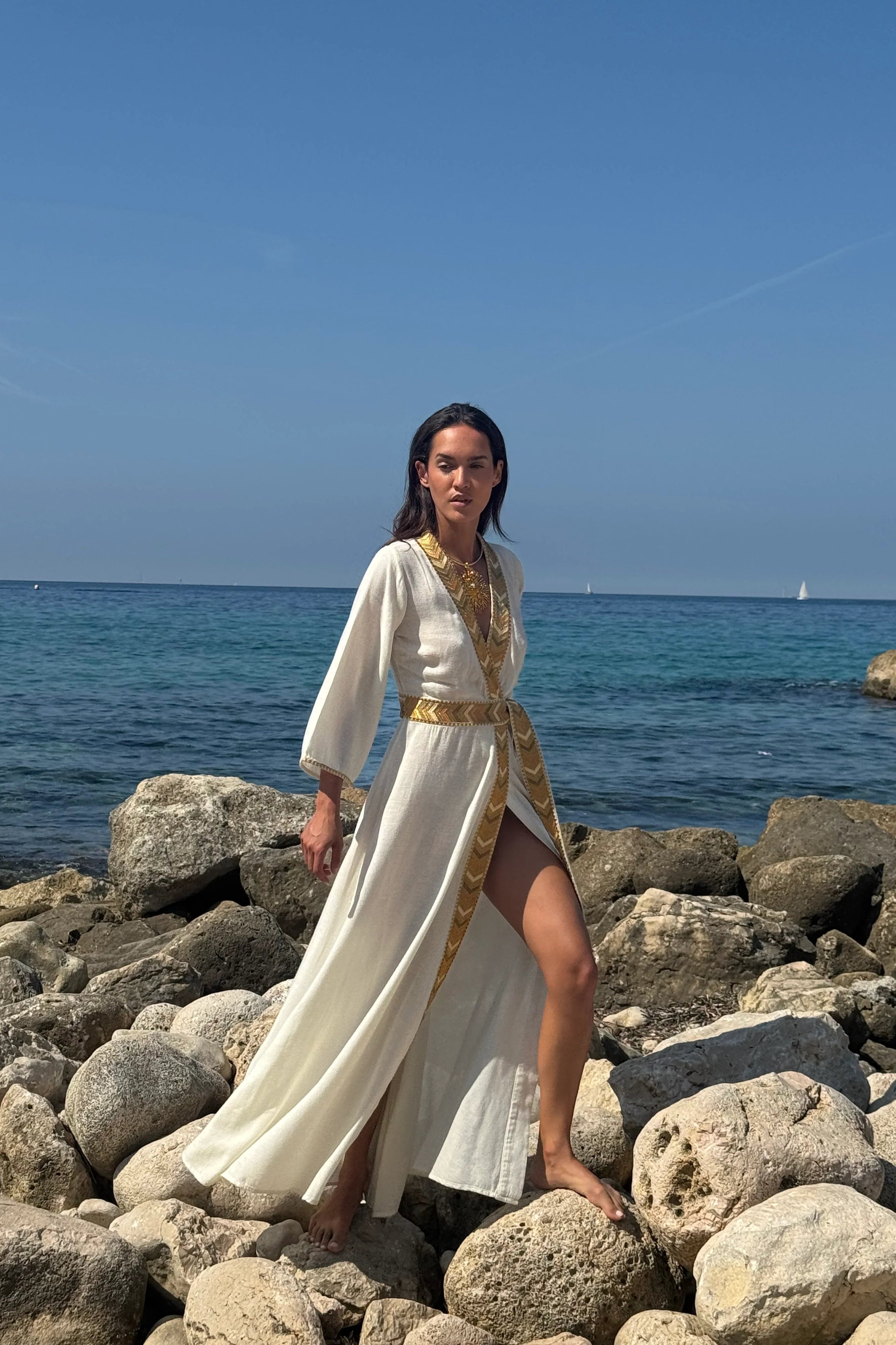 Model wearing the Natacha & Vanessa Livia Maxi Dress with an integrated waist tie and gradient trim on a rocky shoreline.