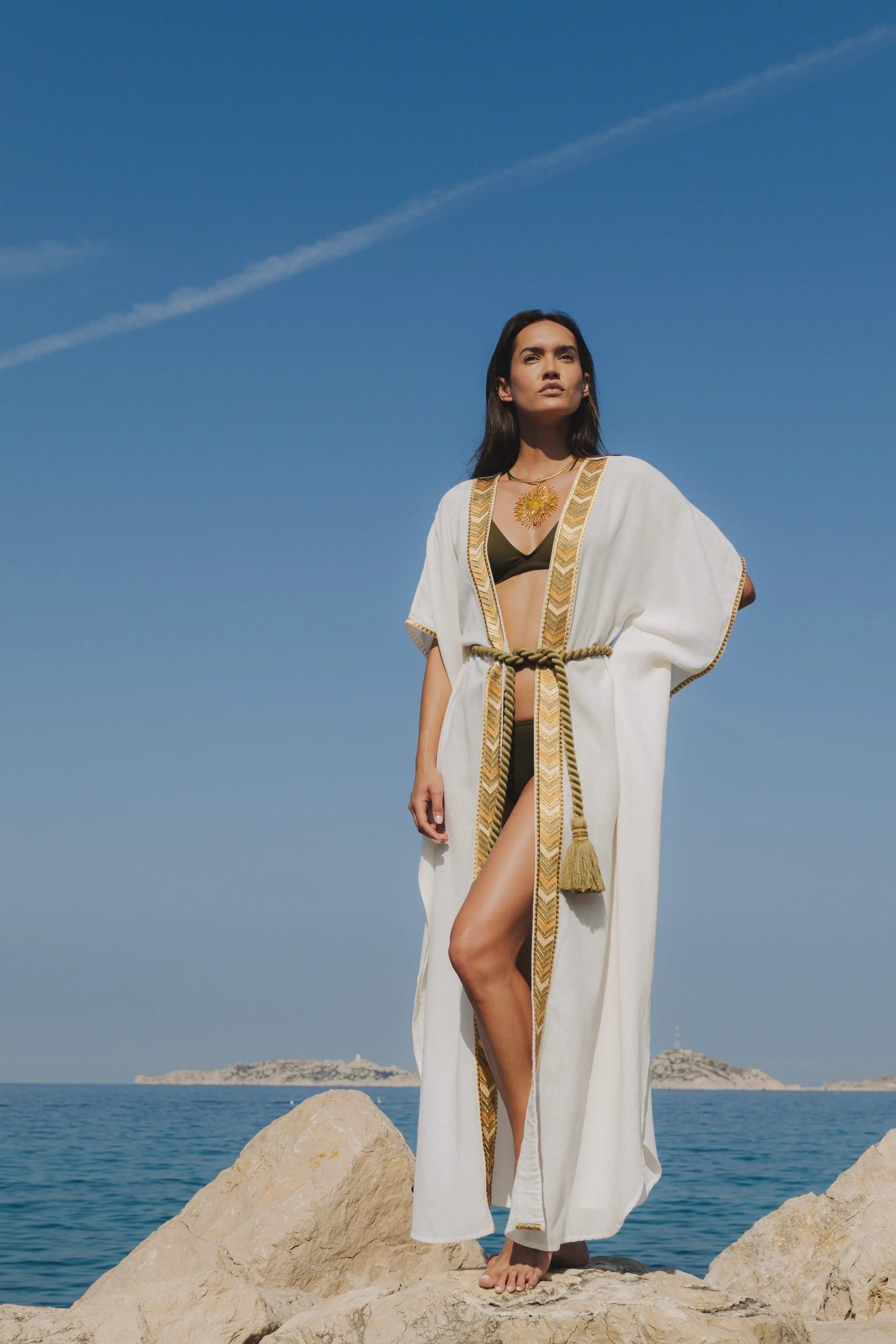 Model wearing the Natacha & Vanessa Ocean Dress in white with gold embroidered trim, kimono sleeves, and a braided rope belt while standing on seaside rocks.