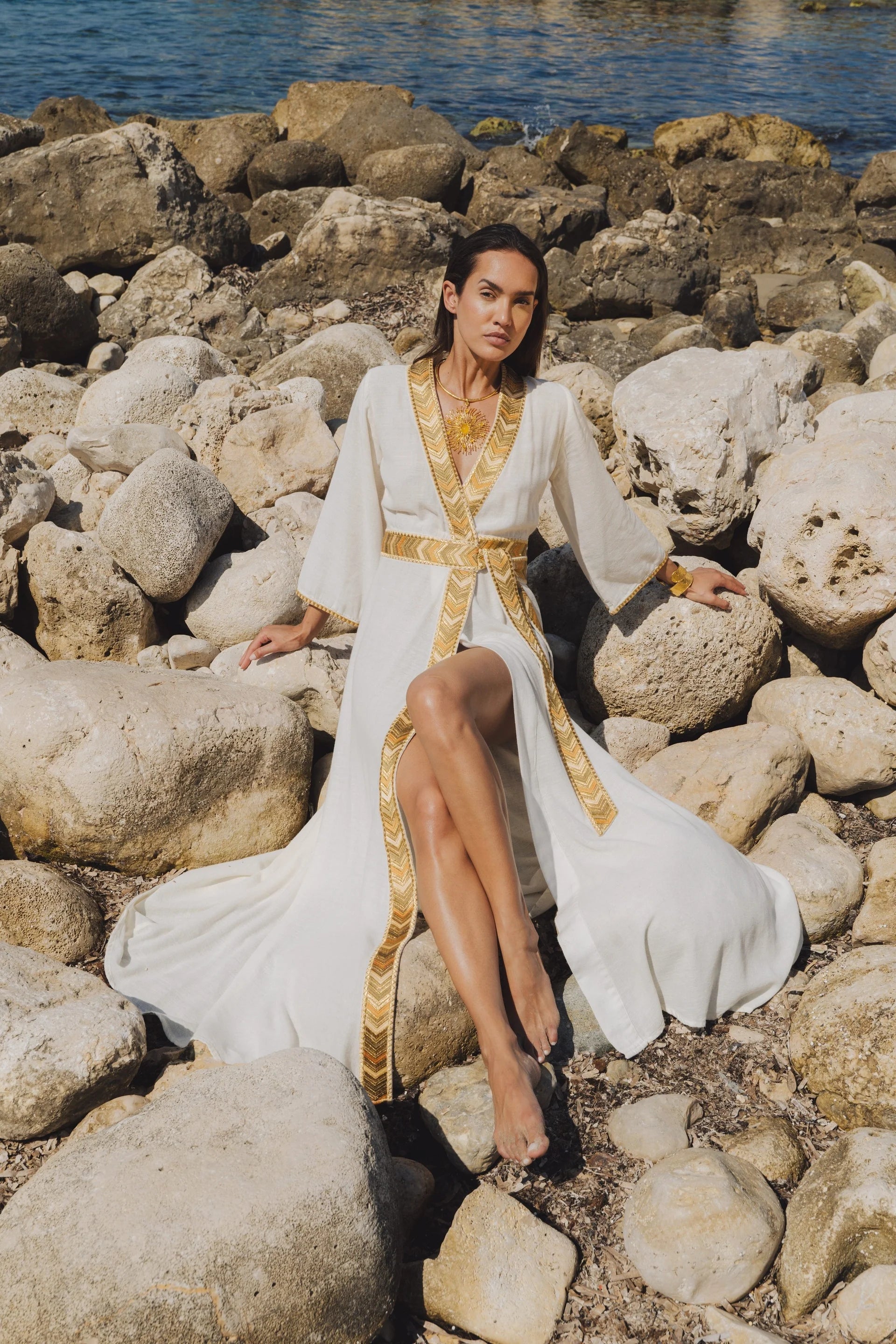 Model wearing the Natacha & Vanessa Livia Maxi Dress in ecru with an ochre and gold gradient trim while seated on seaside rocks.