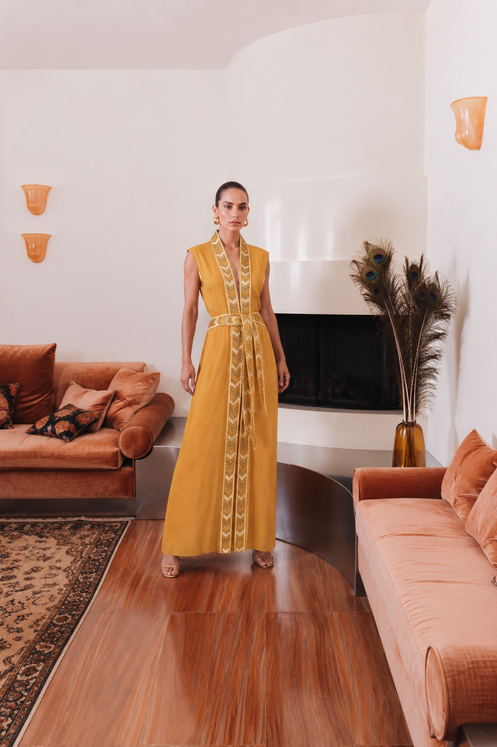 Model wearing the Natacha & Vanessa The Thalie dress in saffron with embroidered trim and braided belt in an elegant interior.