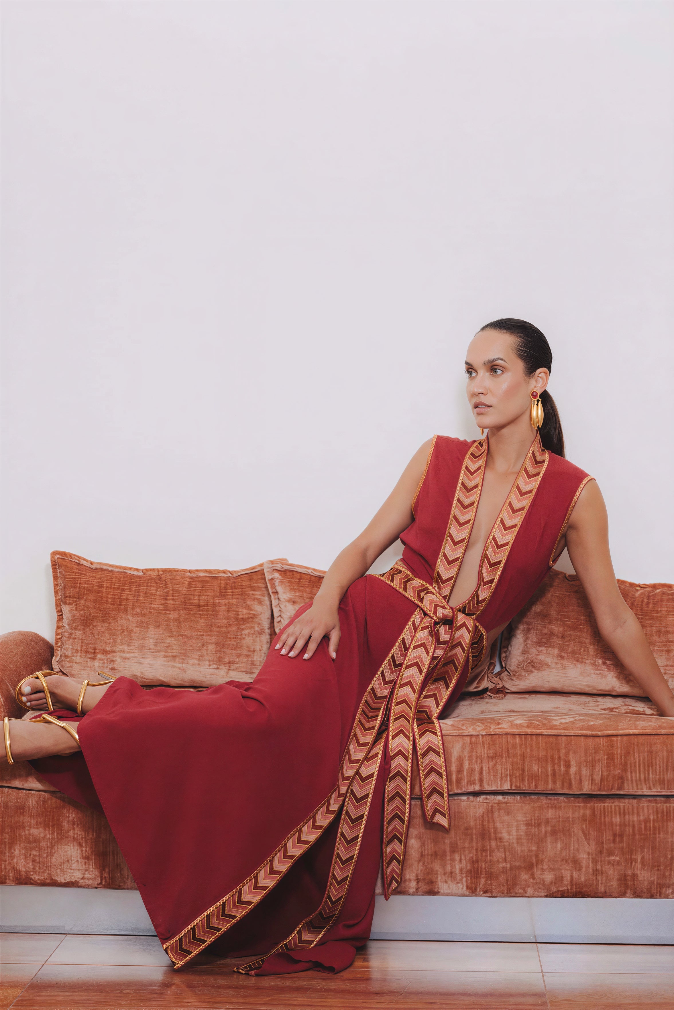 Model seated on a velvet sofa wearing the red Thalie dress featuring a woven border and braided waist belt.